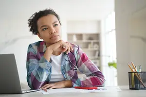 A woman is sitting at a desk with a laptop, papers, and pens, looking thoughtful.