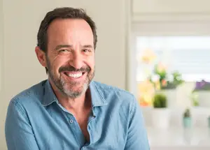 A smiling man with a beard and mustache is wearing a blue shirt and is looking to the right. Behind him, there is a window with a view of a garden with various potted plants and flowers. The man's shadow is cast on the wall to his left.