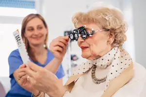 An elderly woman wearing glasses is holding an eye chart while being assisted by a woman in a blue uniform at an optometrist's office.