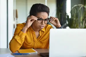 A woman with glasses is sitting at a desk with a laptop and a notebook in an office. She is holding her glasses with both hands and seems to be in a serious expression.