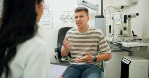 A man wearing a striped shirt and jeans is sitting on a chair and appears to be talking to a woman wearing a white shirt. They are in an indoor setting, which could be a clinic, and there are various pieces of equipment on the desk behind them.