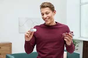 A man in a maroon sweater is holding a pair of glasses and a small lens in his hands while smiling in a living room.