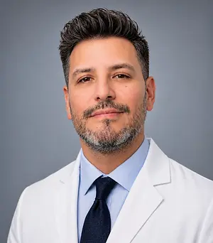 A close-up portrait of a man with short black hair and a beard, wearing a white coat and blue shirt, smiling at the camera.