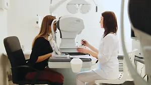 A woman is having her eyes examined by an optometrist in a white coat using a phoropter.