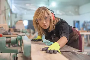 A woman in a workshop, wearing protective eyewear and gloves, bends over to inspect a wooden plank.