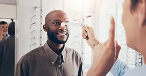 A man wearing a brown shirt and a beard is smiling while a person is holding a ruler in front of his face in a room with a white wall and many glasses hanging on the wall.