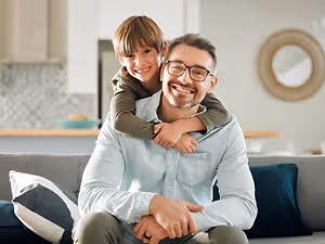 A man is sitting on a couch with a young boy sitting on his shoulders in a living room.