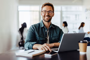 A man is smiling while sitting in front of a laptop on a table with a cup of coffee, a notebook, and a pen.