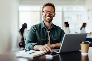 A man is smiling while sitting in front of a laptop on a table with a cup of coffee, a notebook, and a pen.