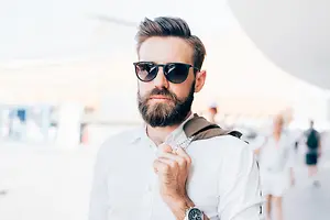 A man wearing a white shirt and a black watch with a beard stands in an outdoor area, adjusting his shirt and looking ahead while a blurry background shows people walking in a hallway.