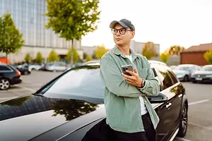 A man in a green jacket is standing next to a black car in a parking lot and looking at his phone.
