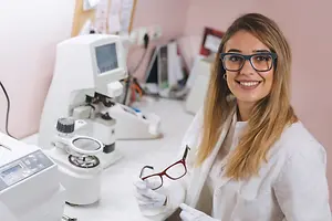 A woman wearing glasses and a lab coat is standing in a room with equipment and smiling