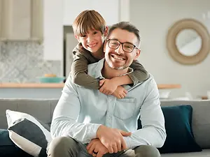 A man wearing glasses is sitting on a couch with a young boy on his back, both smiling