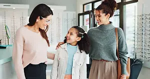 Three people, a mother and two daughters, stand in an optical shop, smiling and looking at each other with the girl wearing glasses.