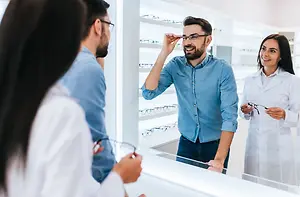 Four individuals, including two women in white lab coats, are standing in a room with a glass wall, discussing glasses with a man who is wearing glasses and holding a pair of glasses in his hand.