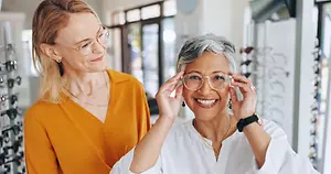 Two women in a glasses shop, one trying on glasses while the other looks on and smiles.