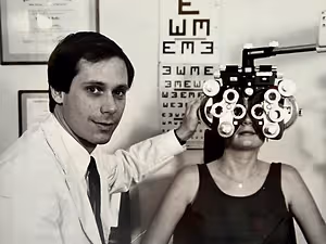 A man in a white coat is checking a woman's eyes with an ophthalmoscope.