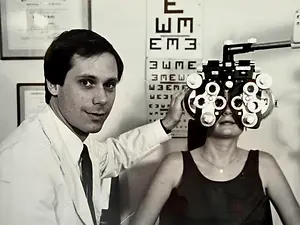 A man in a white coat is checking a woman's eyes with an ophthalmoscope.
