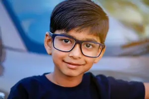 A smiling young boy with glasses posing for a picture in front of a car.