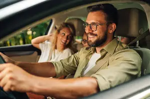 A man wearing glasses is driving a car with a woman sitting in the passenger seat and another person in the backseat, with trees visible in the background.