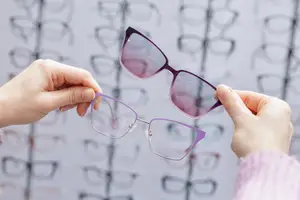 A woman wearing a pink sweater is holding a pair of glasses in front of her face with both hands. Behind her, there is a blurry background of several pairs of glasses with purple frames and black frames with white lenses displayed on a white wall.