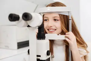 Young girl with long red hair and a white shirt looking into an eye examination machine and smiling