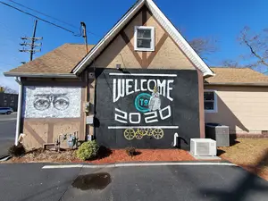 A house with a black wall featuring a mural reading 'Welcome 192020' and a bicycle design, accompanied by plants and an air conditioning unit on the side.
