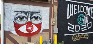 A mural depicting a face wearing a red mask with a star and glasses, next to a black wall with white text reading 'Welcome to 2020' and a blue circle.