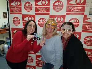Three women in front of a red and white Academy Vision backdrop smiling with 2020 glasses