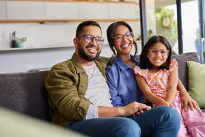 A family of three, including a man, a woman, and a young girl, are sitting on a couch in the living room, all wearing eyeglasses and smiling for a photo.