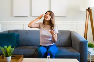 A woman sitting on a couch, holding a thermometer, and looking at it. She is wearing a white top and blue pants. She seems to be checking her temperature.