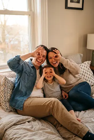 A family of three, a man, a woman, and a young girl, sitting on a bed and smiling while posing for a photo