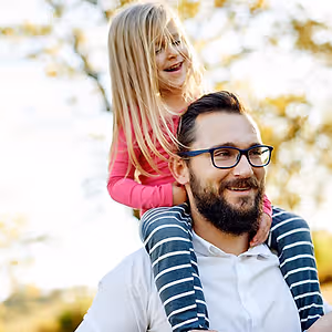 A man with glasses carrying a young girl on his shoulders in a park.