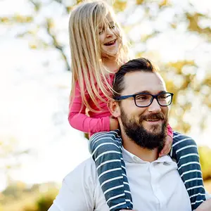 A man with glasses carrying a young girl on his shoulders in a park.