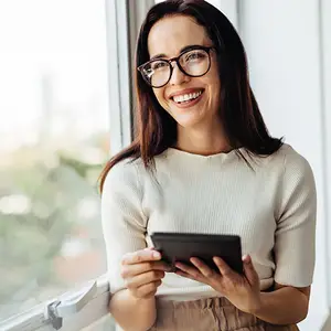 A woman wearing glasses is smiling and holding a tablet in her hands while standing near a window.