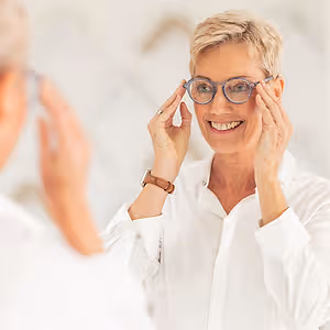 A woman with short hair is smiling while wearing glasses and looking in the mirror. She is holding her glasses in her hands and seems to be trying to adjust them. She is wearing a white shirt and a watch on her left wrist. The other person on the left is holding a pair of glasses and seems to be looking at the woman. Behind the woman is a white wall. 