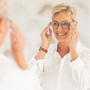 A woman with short hair is smiling while wearing glasses and looking in the mirror. She is holding her glasses in her hands and seems to be trying to adjust them. She is wearing a white shirt and a watch on her left wrist. The other person on the left is holding a pair of glasses and seems to be looking at the woman. Behind the woman is a white wall. 