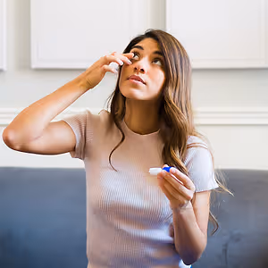 A woman is sitting on a couch and applying eye drops.