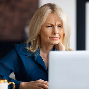 Elderly woman with blonde hair sitting in front of a computer monitor, looking sad or frustrated.