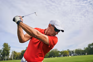 An older man with a red shirt and white hat plays golf on a sunny day