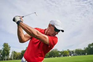 An older man with a red shirt and white hat plays golf on a sunny day