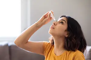 A woman in a yellow shirt is looking up while applying eye drops.