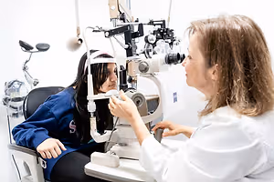 An adult woman wearing a white coat is examining the eyes of a young girl using an ophthalmoscope in a white room.