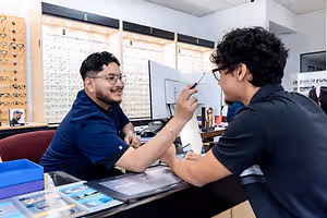 Two men in an optical shop, one with glasses pointing at a monitor while the other holds a pen, both smiling and focused on the task at hand.