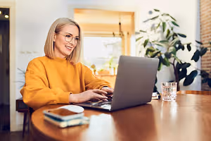 A woman wearing glasses sitting at a table using a laptop while smiling