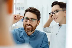 Two young men standing in front of a mirror, one wearing glasses and adjusting his, while the other holds a frame and smiles, both wearing white shirts and eyeglasses in an optical shop.