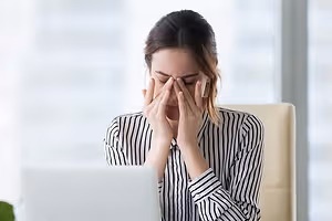 A woman sitting at a desk in front of a laptop, holding her head in her hands, looking stressed.