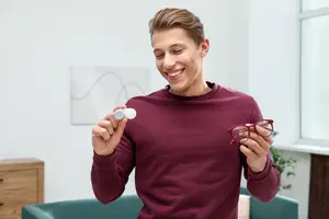 A young man wearing a maroon long-sleeve shirt and holding a pair of glasses and a white contact lens in his hands and smiling