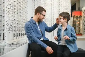 A man and a boy sitting on a bench in an optical store, looking at a glass case of eyeglasses