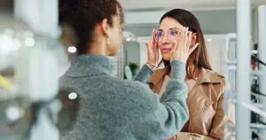 A woman wearing glasses is holding her face in her hands while looking in a mirror. Another woman is standing behind her. Both women are wearing earrings and bracelets.